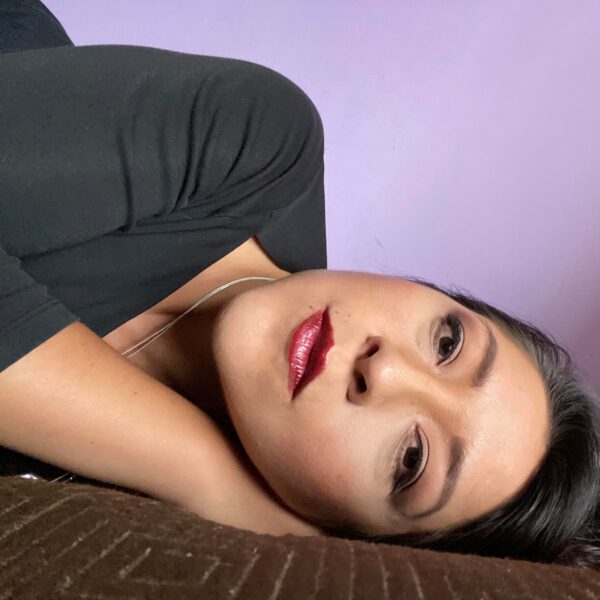 Maritza recostada de lado, con expresión introspectiva, labios rojos y fondo lila, retratando un momento de pausa, reflexión y fortaleza interior.