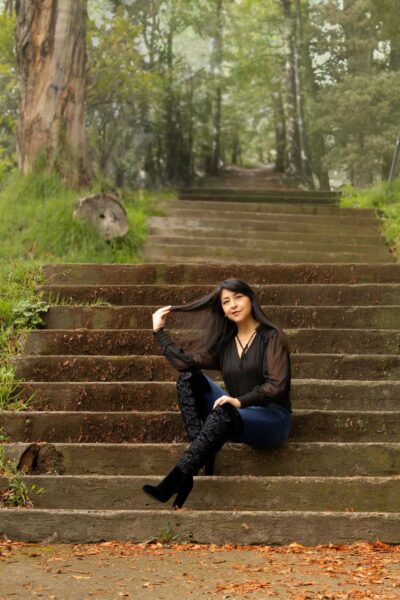 Maritza sentada en unas escalinatas de piedra en un parque rodeado de árboles, con expresión tranquila y gesto relajado, peinando suavemente su cabello.