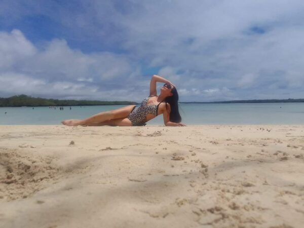 Maritza recostada sobre la arena en traje de baño, disfrutando del sol y el mar en una playa de aguas tranquilas, expresión relajada y empoderada.