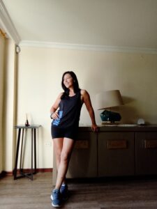 Mujer de pie junto a una mesa, vestida con ropa deportiva, sonriendo con confianza en un ambiente interior iluminado.